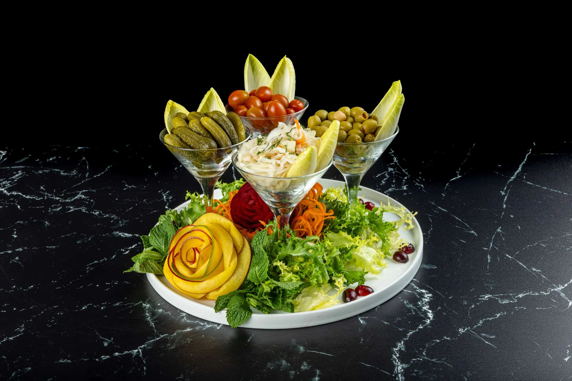 Copas de encurtidos, aceitunas y ensalada de col sobre una fuente blanca con ensalada mixta y una rosa de calabacín amarillo
