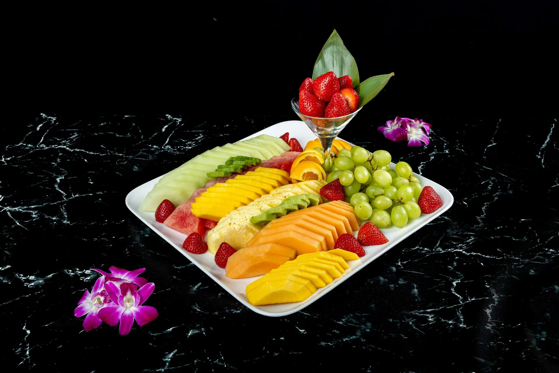Una fuente blanca de melón, mango, kiwi, uvas y fresas en rodajas con una orquídea