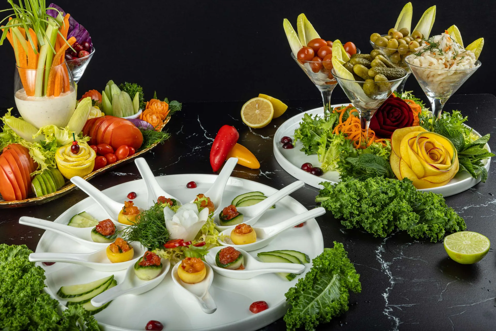 Una torre de bastones de zanahoria, pepino y pimiento con hojas de endivia, y delante una fuente redonda de canapés en cuchara