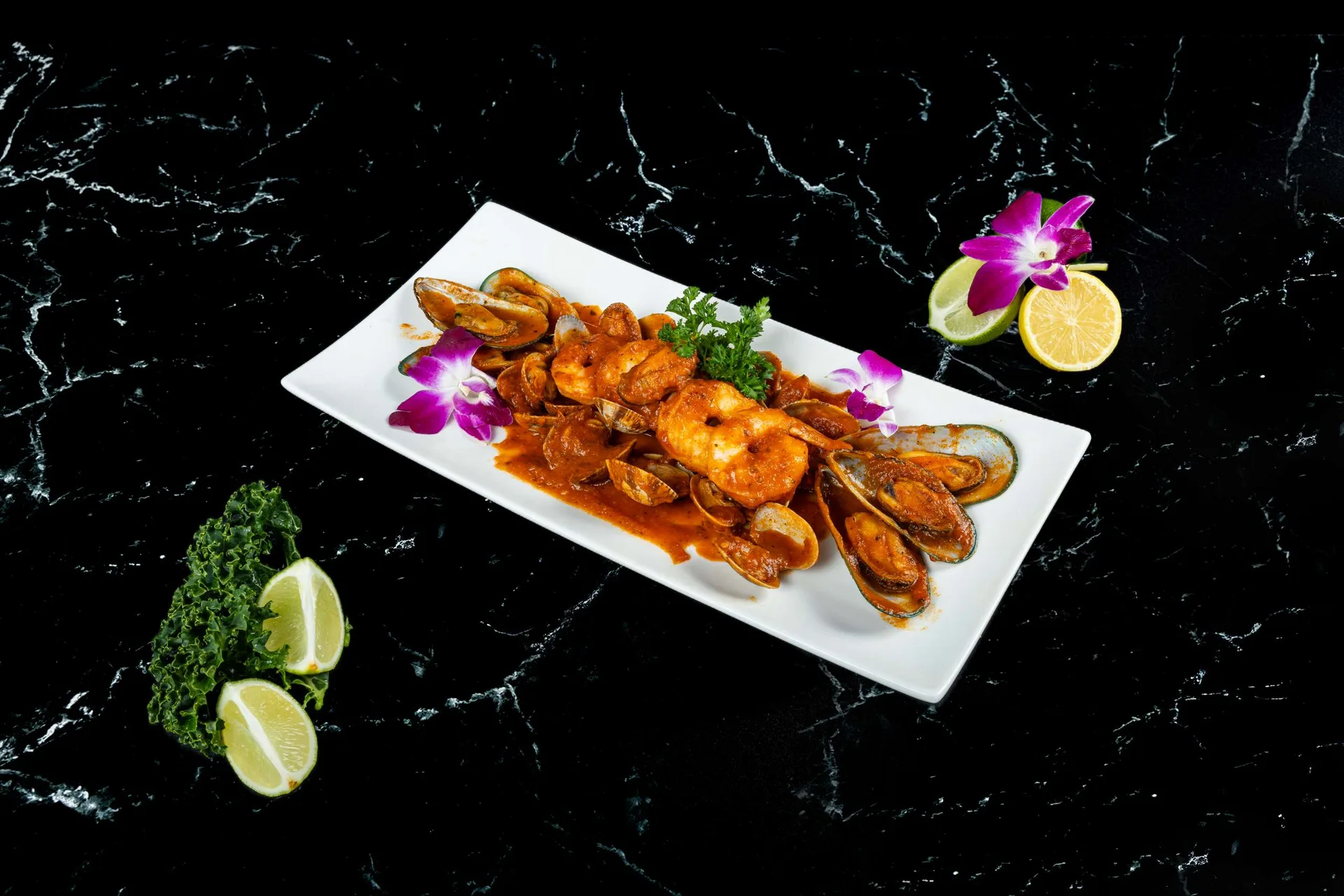 Gambas y mejillones en salsa de tomate en una fuente rectangular con gajos de lima y una orquídea