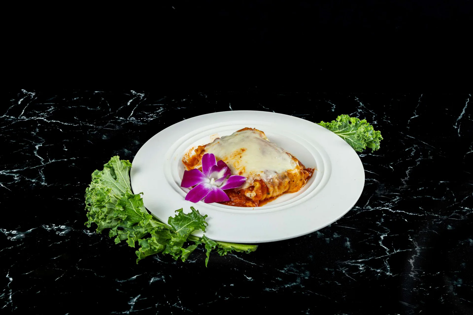 Pasta al horno en salsa de tomate con queso fundido en un plato blanco redondo con col rizada y una orquídea