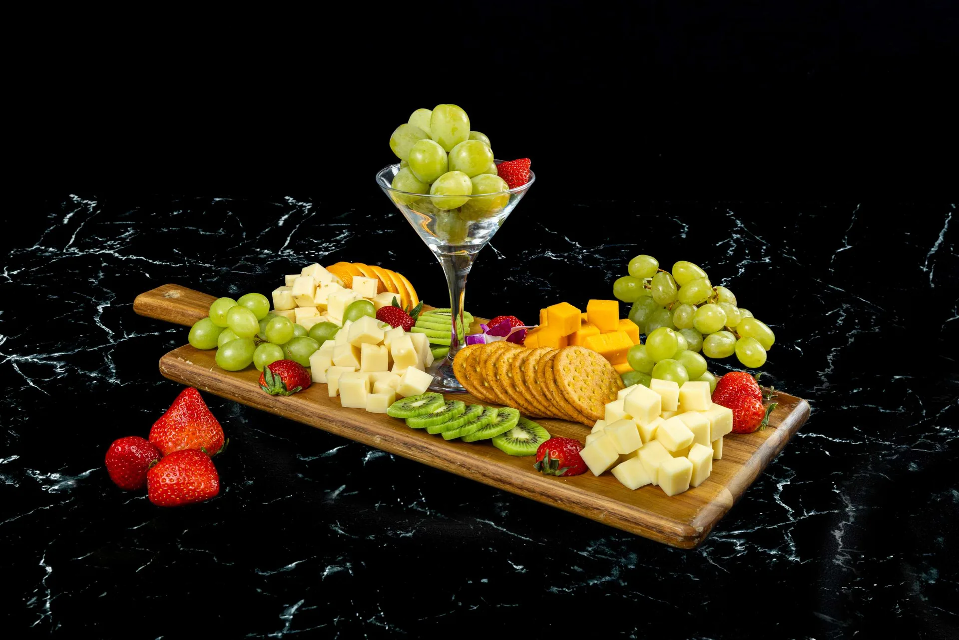 Una tabla de madera con quesos en dados, galletas, uvas y kiwi, y una copa de uvas