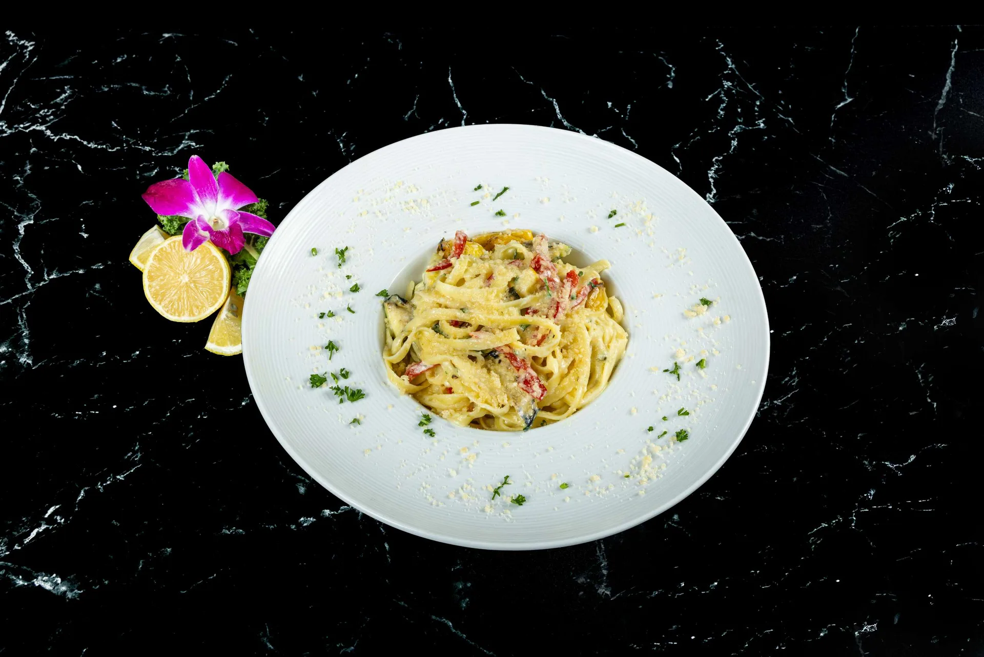 Fettuccine alfredo en un plato blanco redondo con perejil, limón y una orquídea