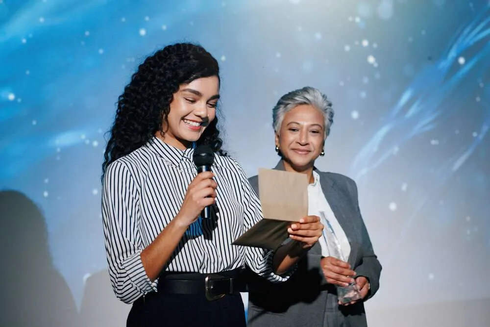 Announcement, awards ceremony and presentation with business women on stage for corporate event. Microphone, prize giving and reading winner with employee people in auditorium or convention center.