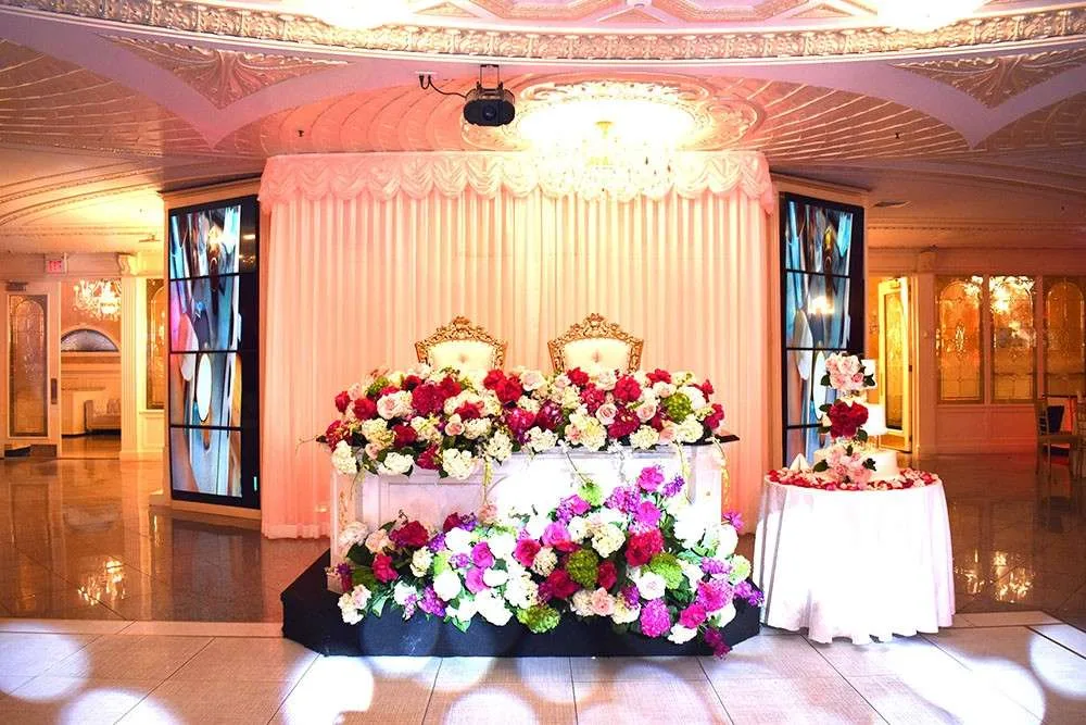 Elegant wedding sweetheart table adorned with vibrant red, pink, and white floral arrangements. Ornate golden chairs sit behind the table, with a chandelier overhead, creating a romantic and luxurious atmosphere.