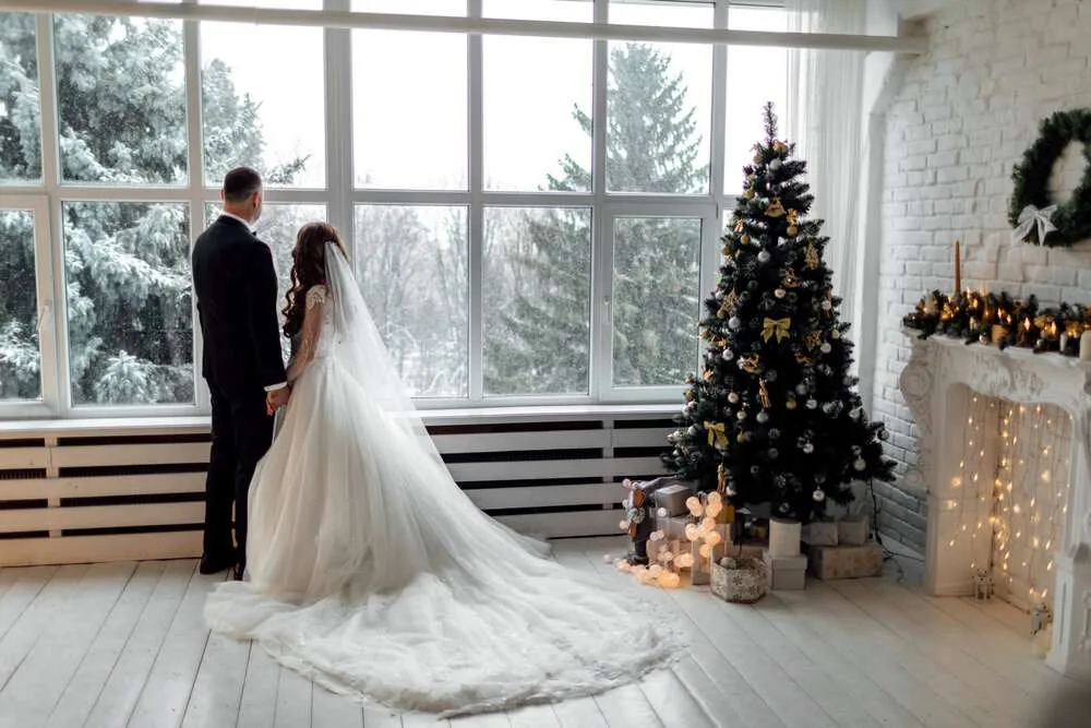 Young couple in love bride and groom posing in studio on background decorated with Christmas tree in their wedding day at Christmas near the large panoramic window. copy space