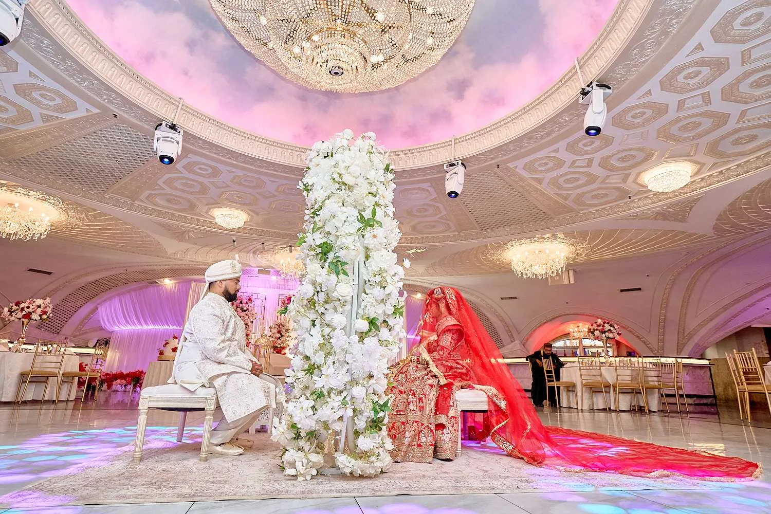 El novio sentado en un mandap de flores blancas mientras el largo dupatta rojo de la novia se extiende por la pista