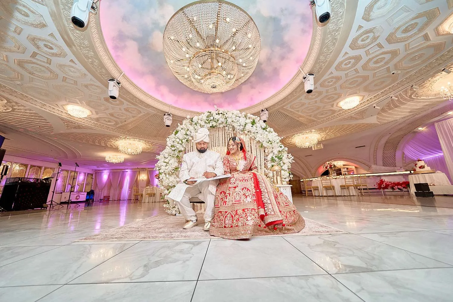 La pareja sentada junta en el mandap, la novia con lehenga roja y el novio de crema, bajo la cúpula de nubes