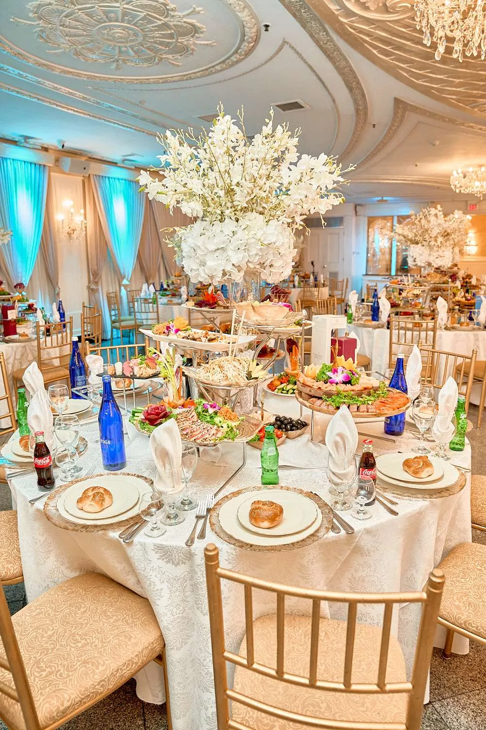 Una mesa de banquete con panecillos de jalá en cada cubierto y bandejas escalonadas de fiambres y ensaladas