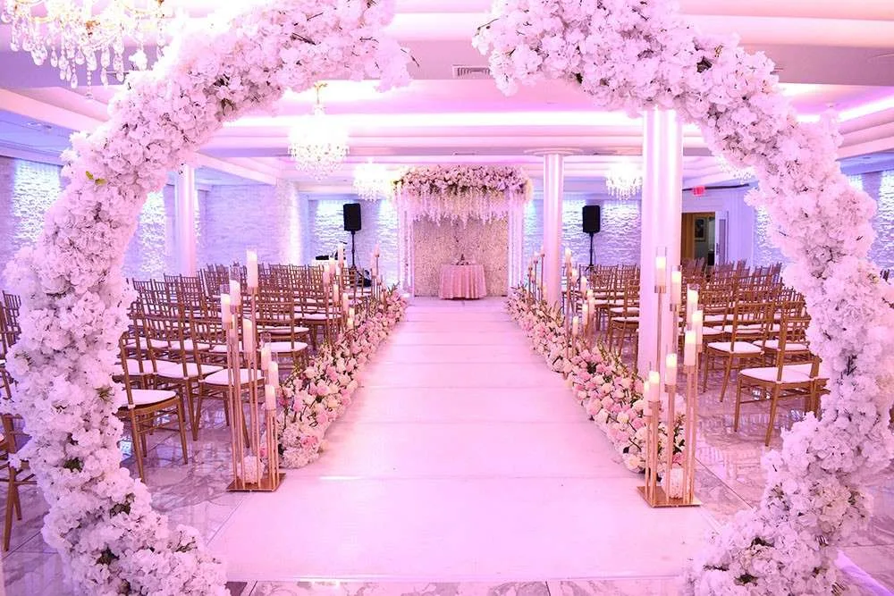 Un pasillo de ceremonia bajo un dosel de flores rosadas, con columnas de velas y sillas doradas