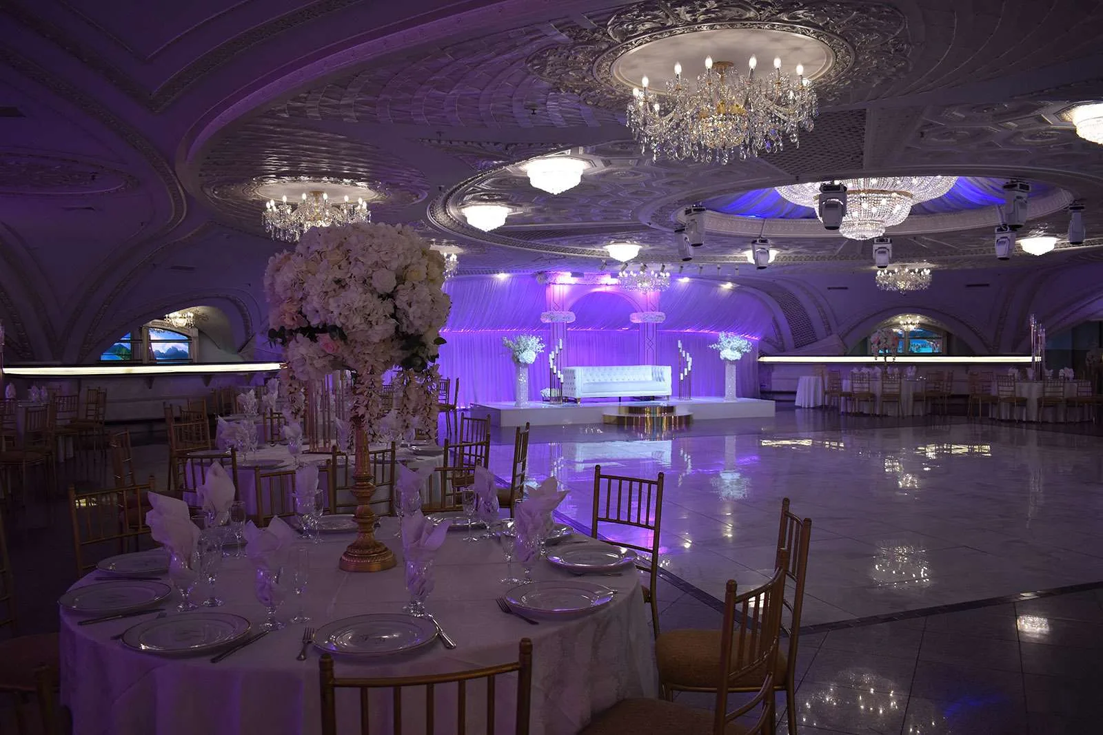 El salón con un escenario blanco, altos centros de mesa blancos y sillas doradas con luz morada