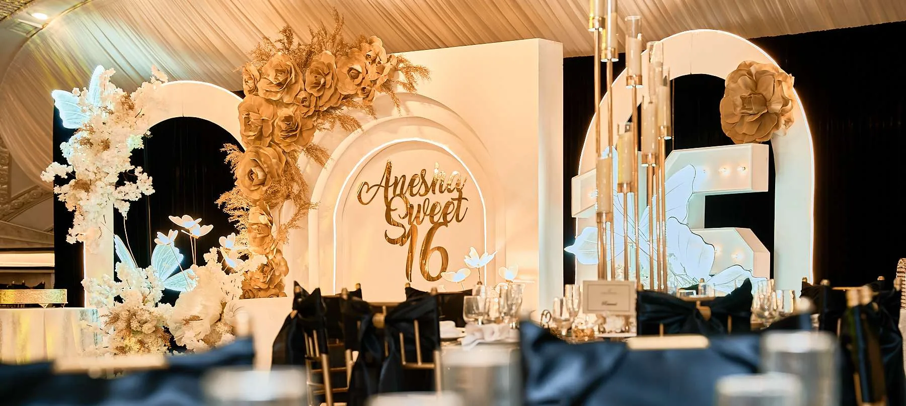 Un letrero dorado iluminado de Sweet 16 en un arco blanco de rosas de papel, con sillas de funda negra