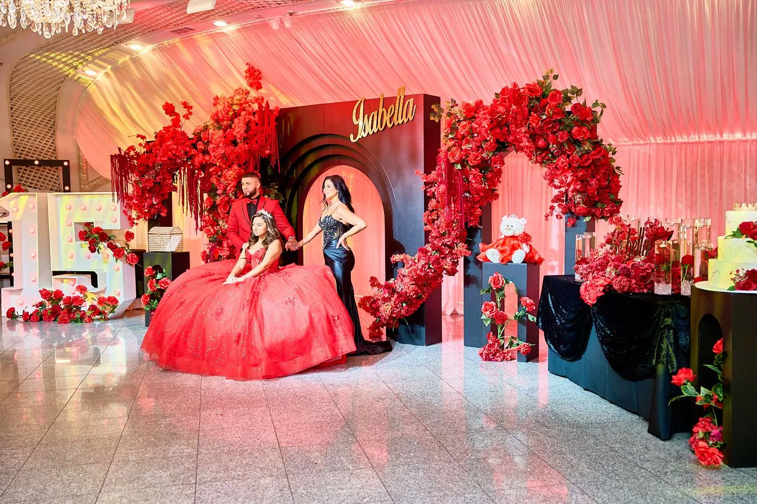 La homenajeada con vestido rojo junto a su familia ante un arco de rosas rojas