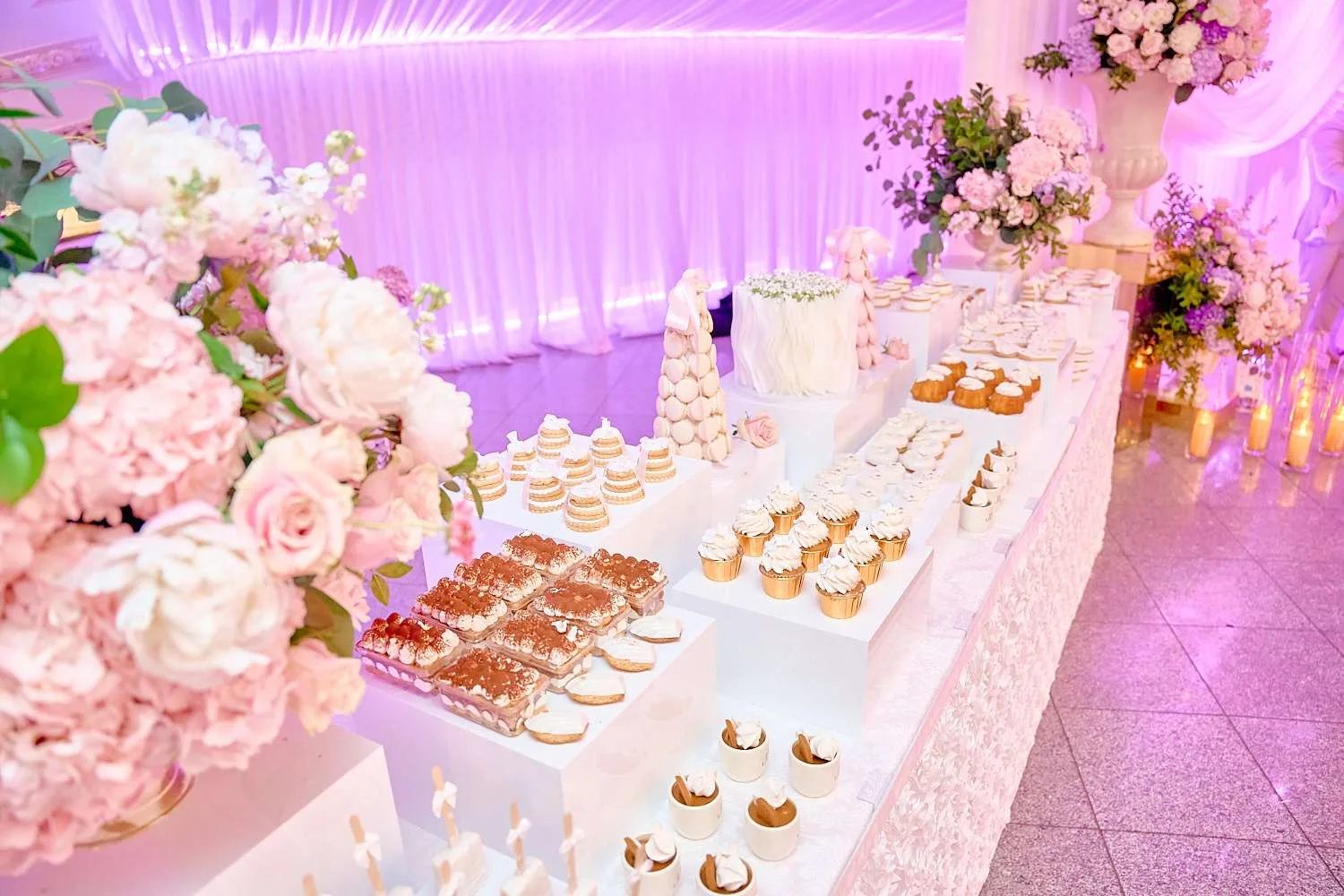 Una mesa de postres con cupcakes, macarons y tartaletas sobre bases blancas, rodeada de peonías y rosas rosadas