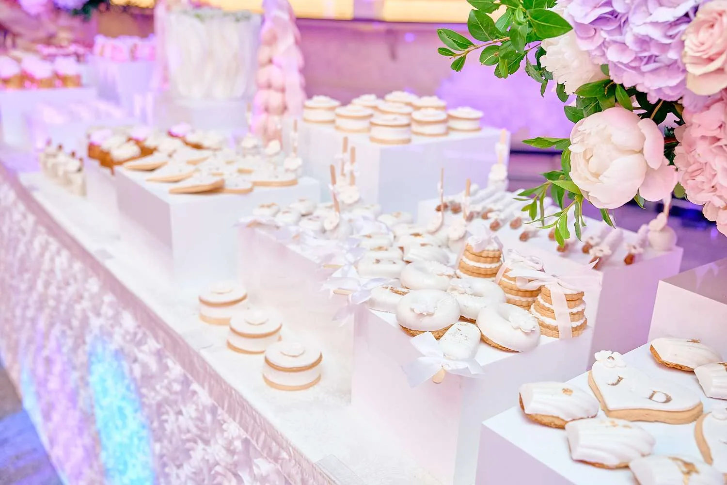 Una mesa de postres blanca con galletas glaseadas, donas y cupcakes junto a una torre de algodón de azúcar