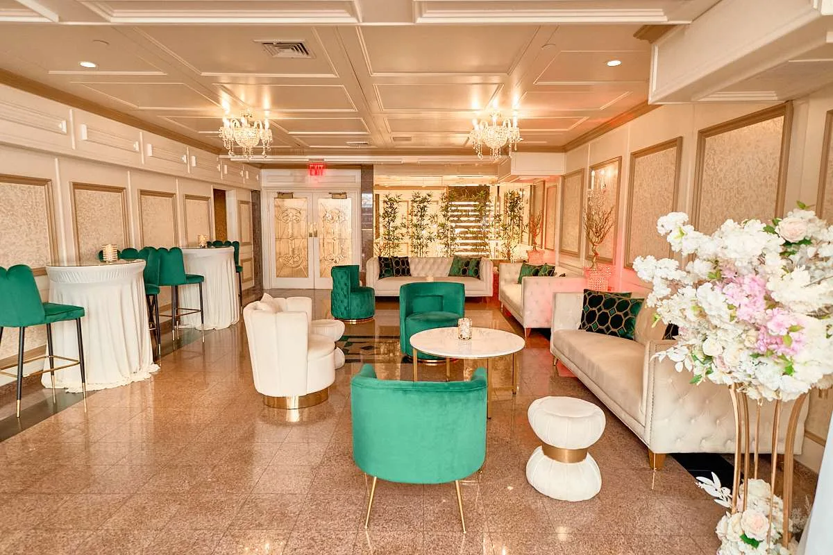 Un salón de cóctel con sillas de terciopelo turquesa y mesas altas blancas, un panel floral dorado y flores blancas