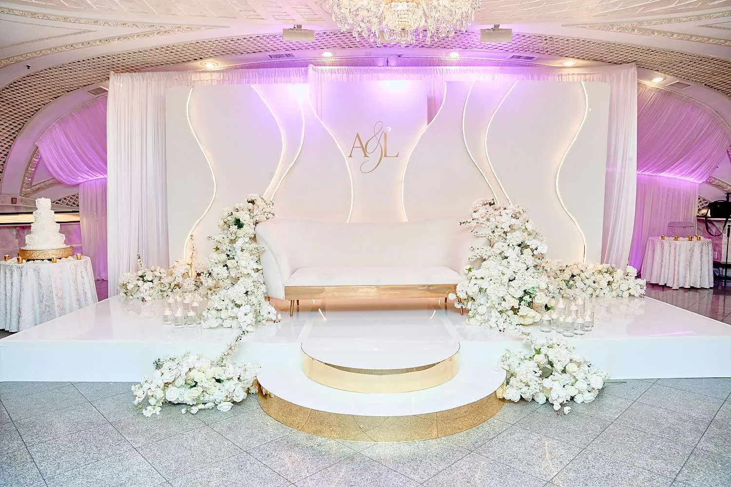 Una tarima elevada para los novios con un sofá blanco curvo, un fondo de cortinaje con monograma y macizos de flores blancas