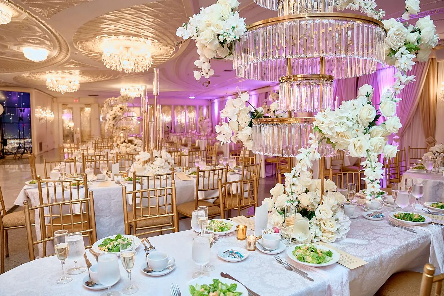 Una mesa larga con mantel blanco y platos ribeteados en oro bajo una estructura suspendida de varillas de cristal y orquídeas blancas