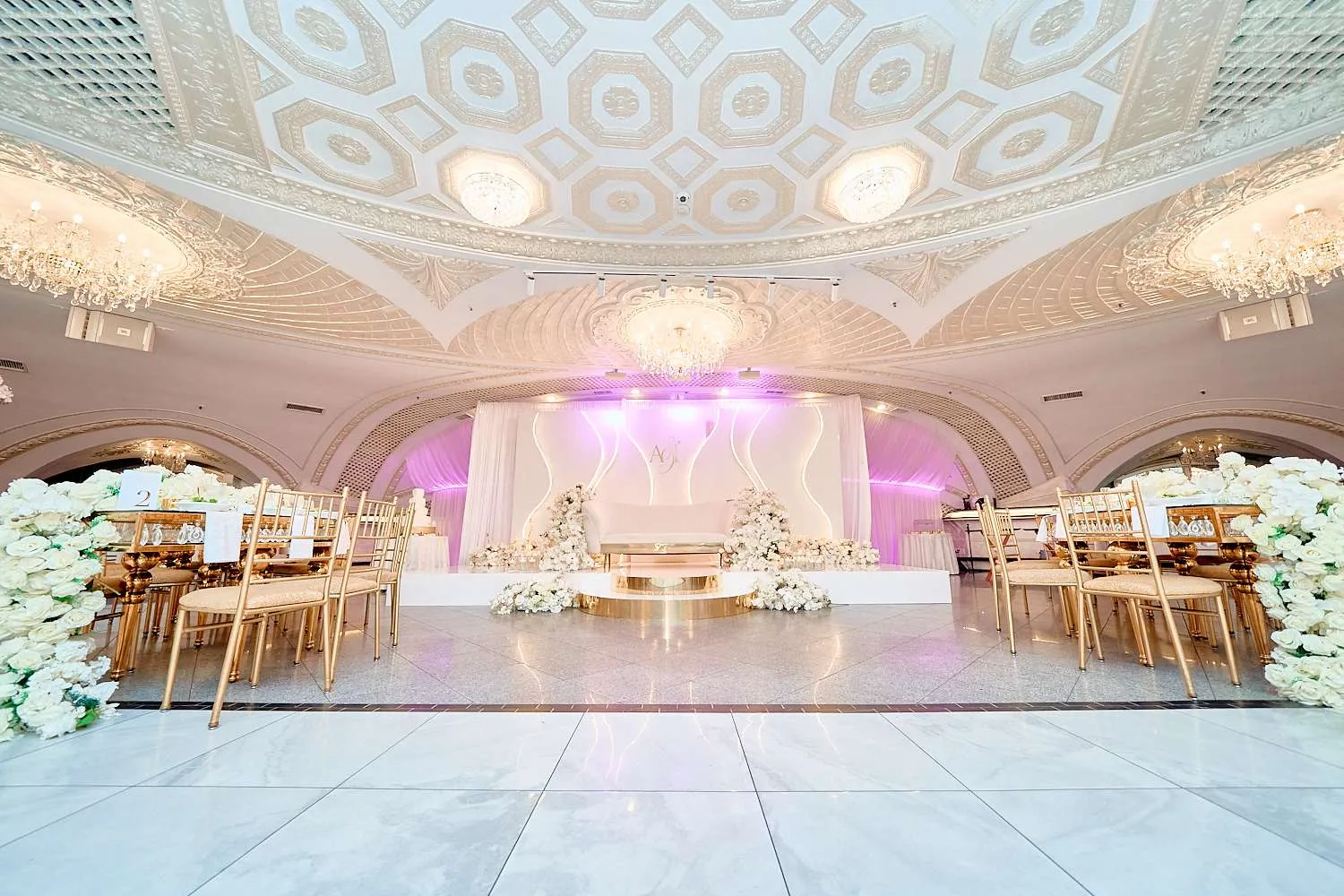 El salón dispuesto con un escenario blanco y macizos florales bajo un techo geométrico dorado