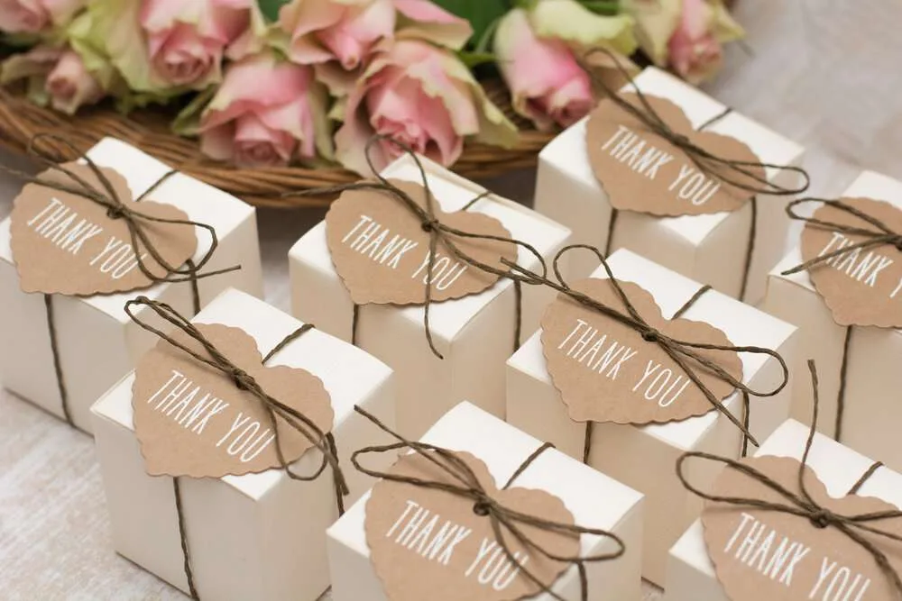 Filas de cajitas de recuerdo atadas con cordel y etiquetas de agradecimiento, junto a un cesto de rosas rosadas