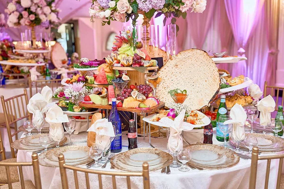 Una mesa servida con bajoplatos dorados y servilletas plegadas ante una bandeja escalonada de fruta, ensaladas y un gran pan redondo