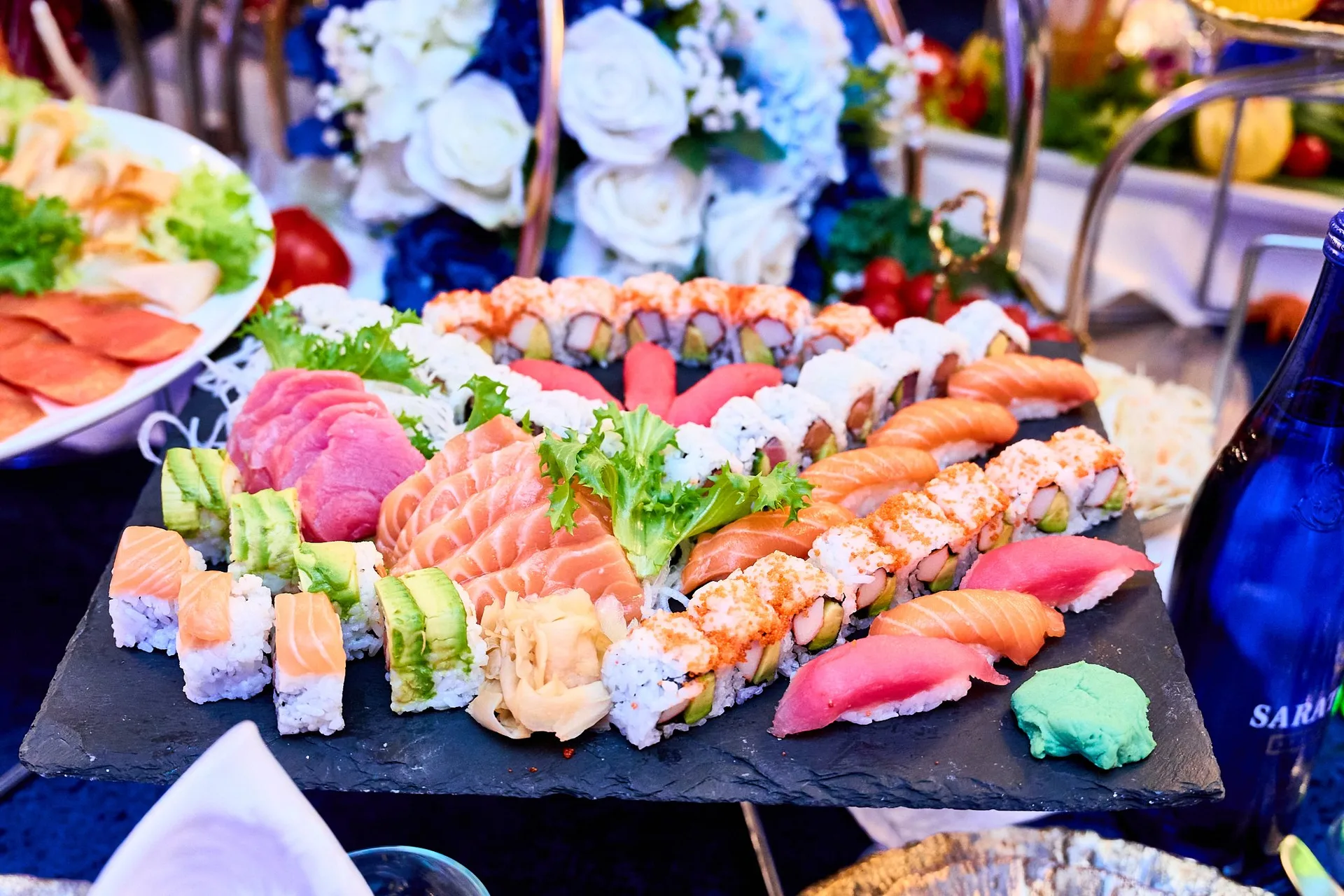 Una tabla de pizarra con sashimi de salmón y atún y rollos de sushi, adornada con rosas blancas