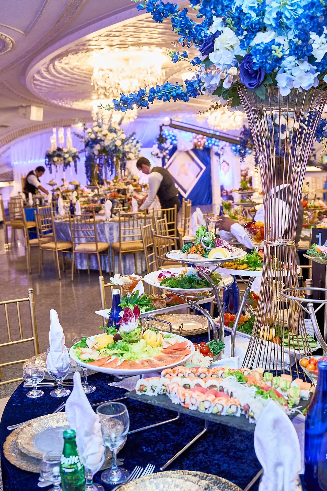 Mesas con mantel azul, bajoplatos de plata, fuentes frías y un alto jarrón de flores azules y blancas