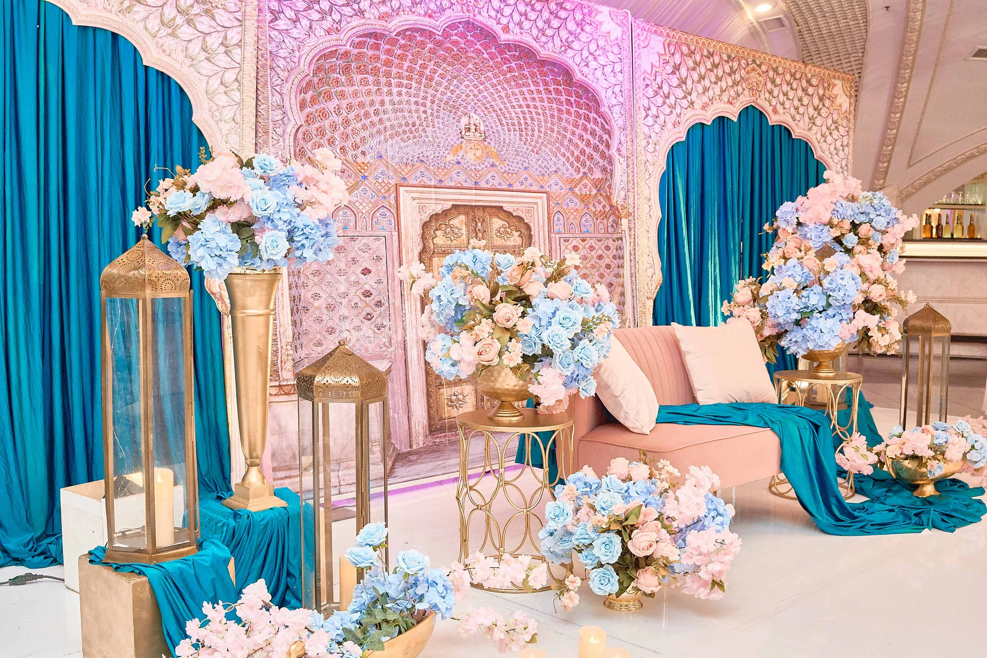 Un escenario lounge en tono rosado ante un fondo de arco mogol tallado, con cortinas turquesa, faroles dorados y hortensias azules