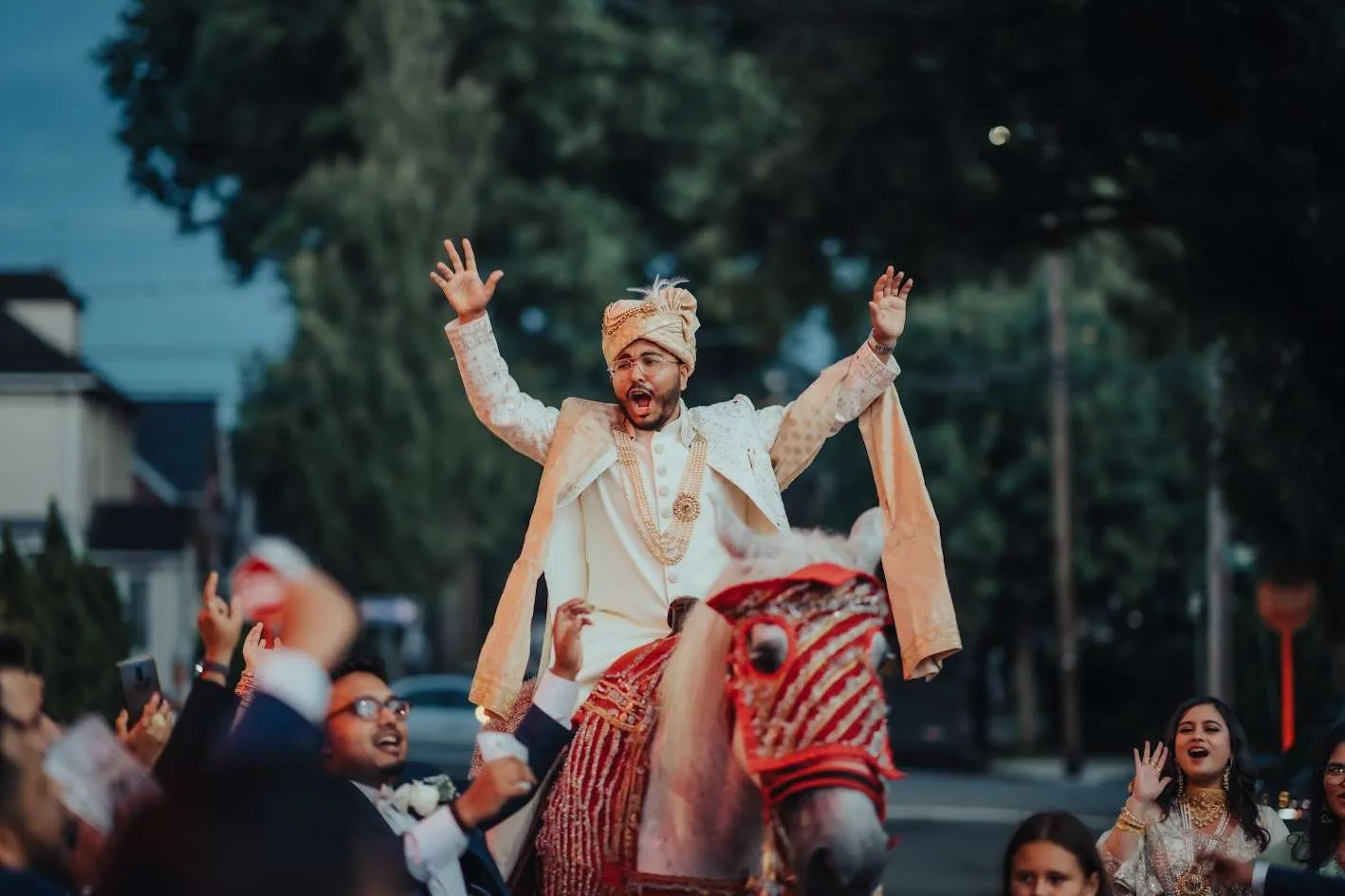 Un novio con sherwani color crema y turbante alza los brazos sobre un caballo blanco engalanado, rodeado de invitados que bailan