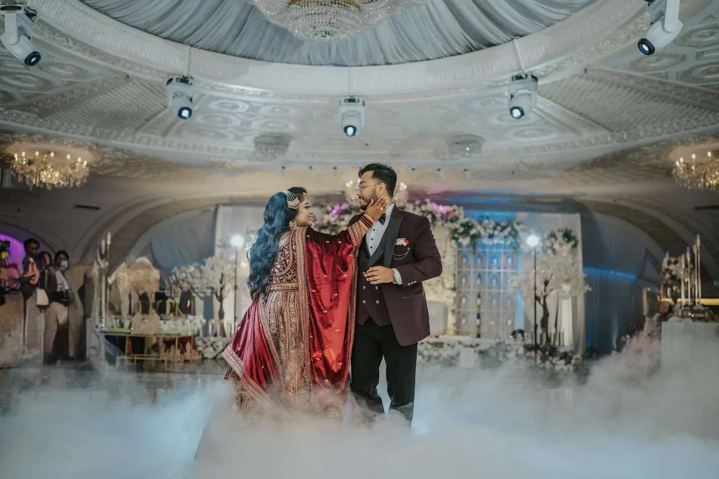 La pareja baila entre una niebla baja de hielo seco, la novia con lehenga rojo intenso y el novio de burdeos