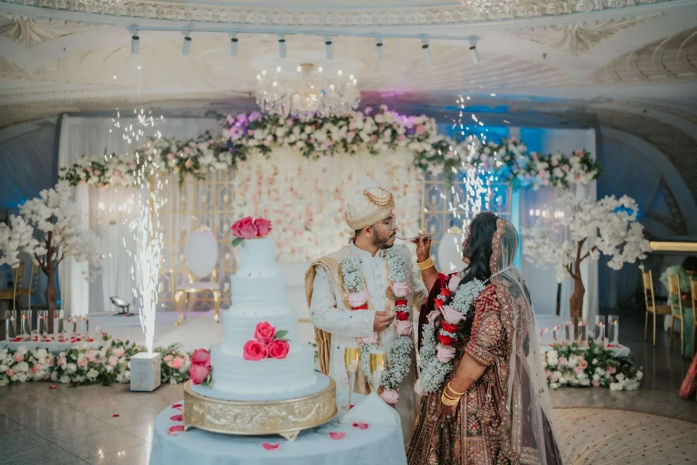 Una pareja corta un pastel de pisos blanco entre fuentes de bengalas, con árboles de flores blancas detrás