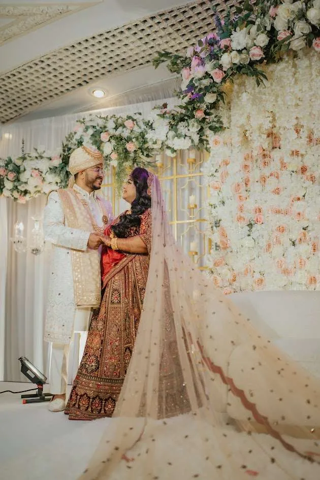 Una pareja bajo un arco floral blanco y rosado, el novio con sherwani crema y la novia de rojo y dorado