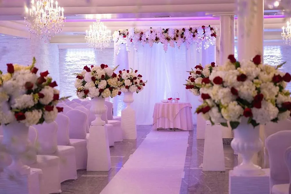 Arreglos de rosas rojas y blancas en urnas blancas a ambos lados del pasillo, ante un fondo de tela blanca con una cenefa floral