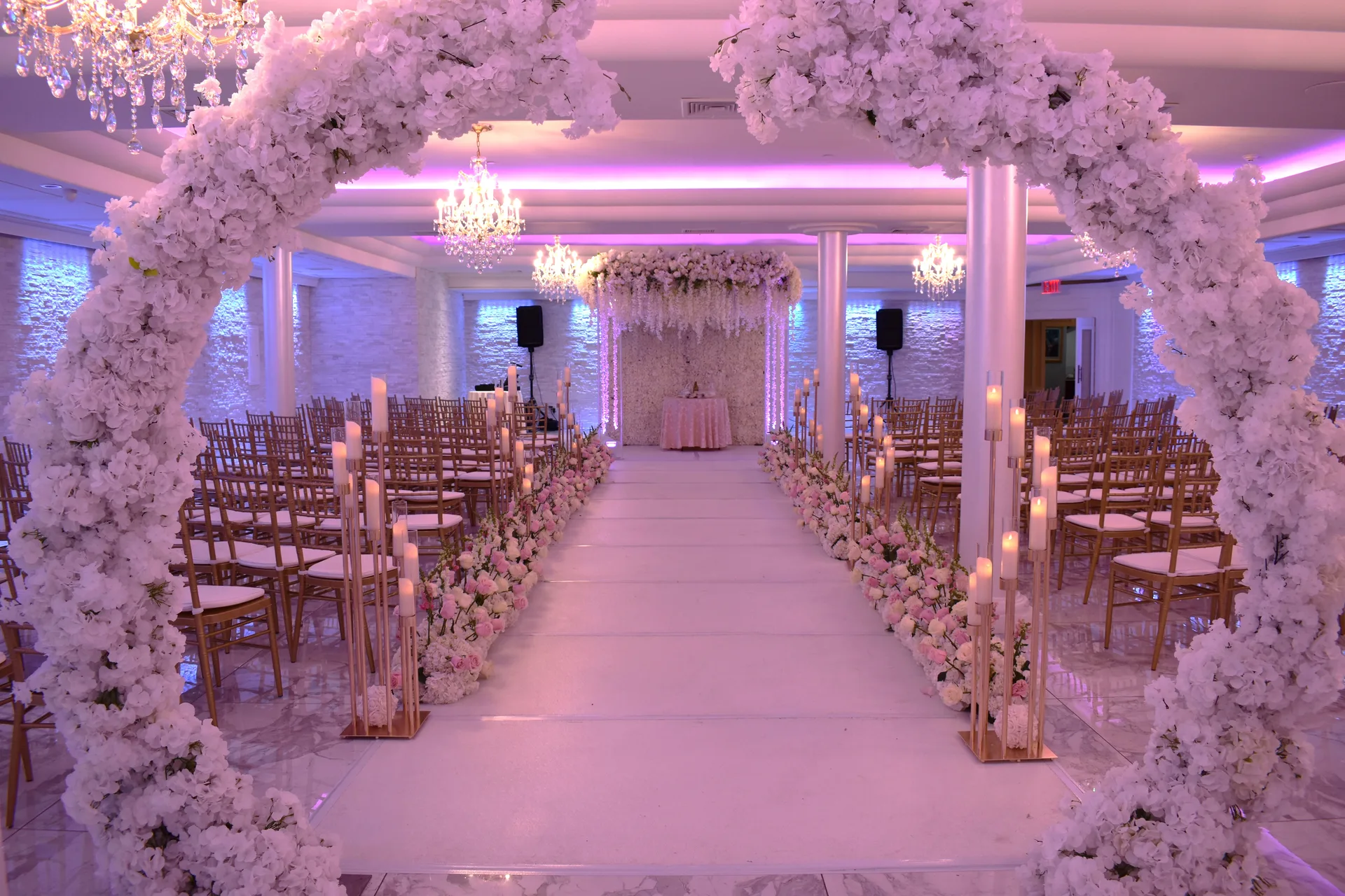 Un arco de flores blancas enmarca el pasillo de la ceremonia, con sillas doradas en filas, columnas de velas y un dosel colgante de glicinas al fondo