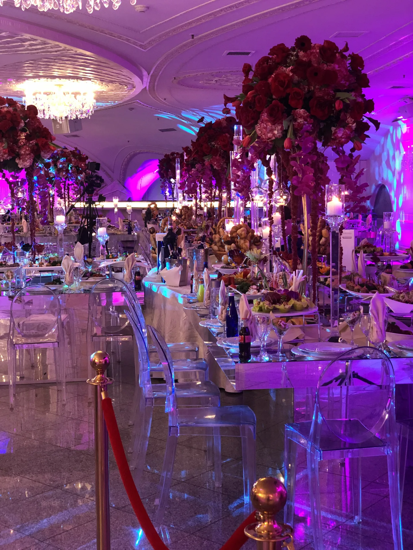 Una mesa larga de banquete bajo altísimos arreglos de rosas rojo oscuro, con sillas de acrílico transparente y bandejas colmadas
