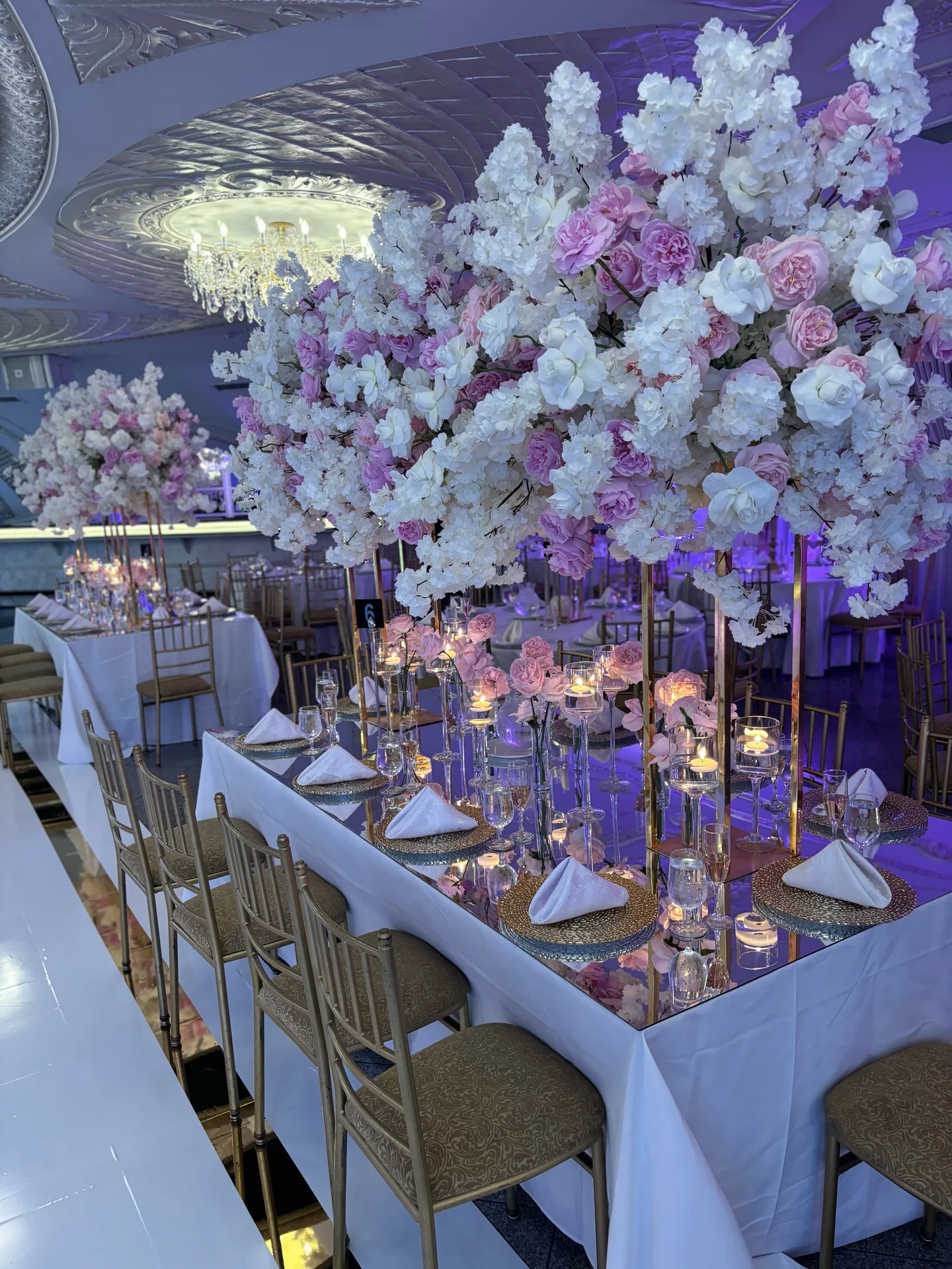 Una mesa de banquete con tablero de espejo flanqueada por árboles de flores blancas y rosadas, sillas chiavari doradas y vajilla ribeteada en oro