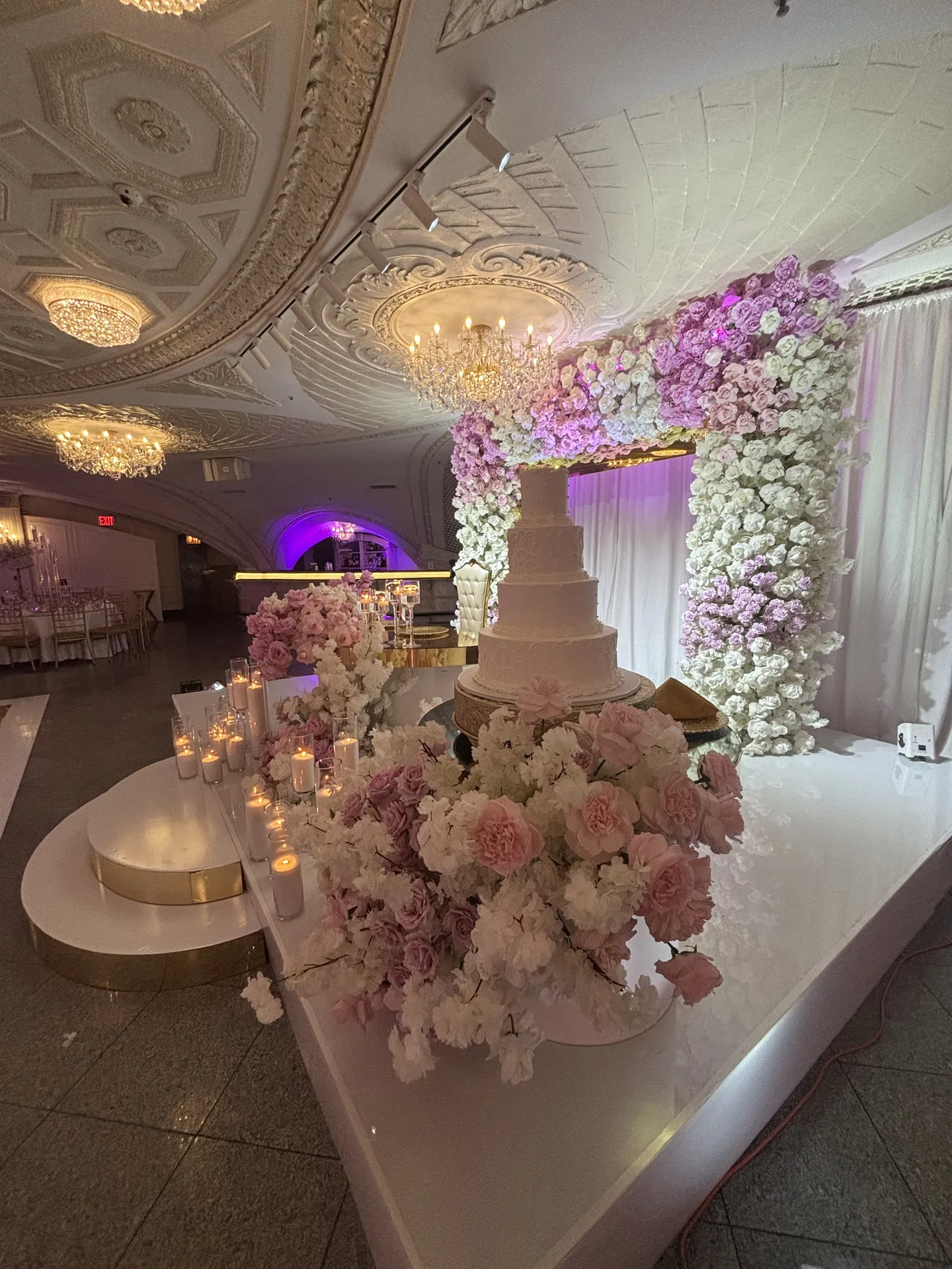 Un pastel blanco de cinco pisos sobre una plataforma elevada dentro de un arco de rosas rosas y blancas, con velas a lo largo del borde