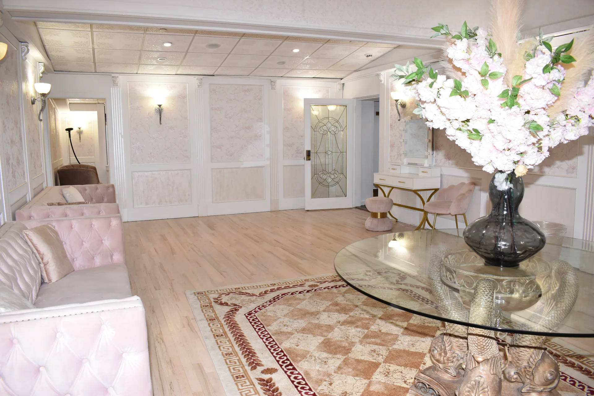 Un sofá capitoné rosado junto a la pared con boiserie de la suite, al lado de una puerta emplomada y un pequeño tocador con silla rosa