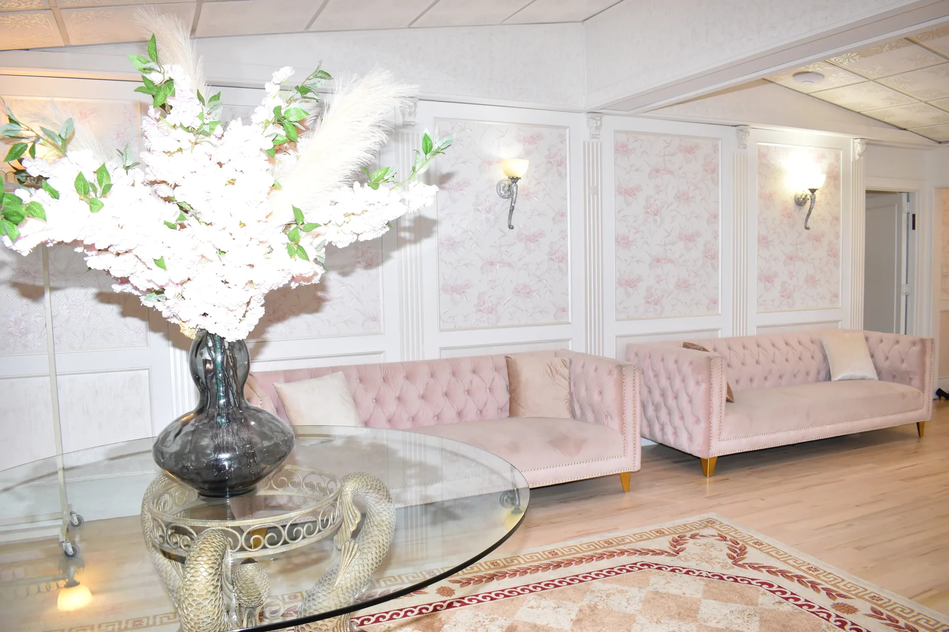 Dos largos sofás de terciopelo rosado bajo apliques y paneles de papel pintado floral, con una mesa de cristal y un arreglo floral delante