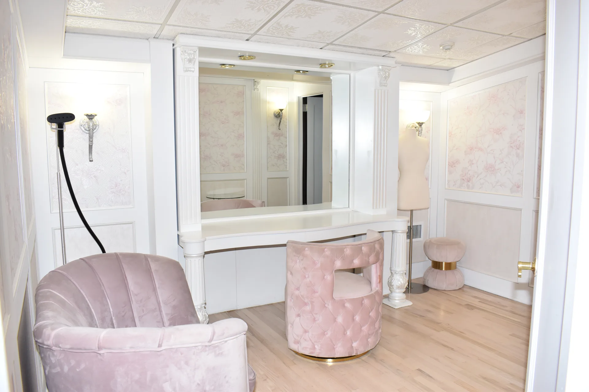 El rincón del tocador de la suite: un espejo ancho entre columnas blancas estriadas, una silla capitoné rosa, una butaca concha y un maniquí
